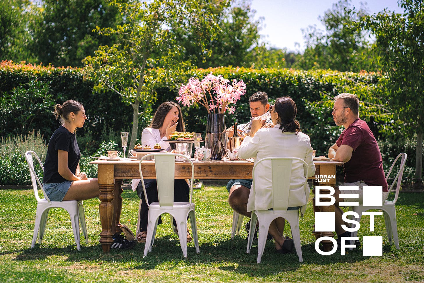 A group of people sit around a high tea table setting in a garden.